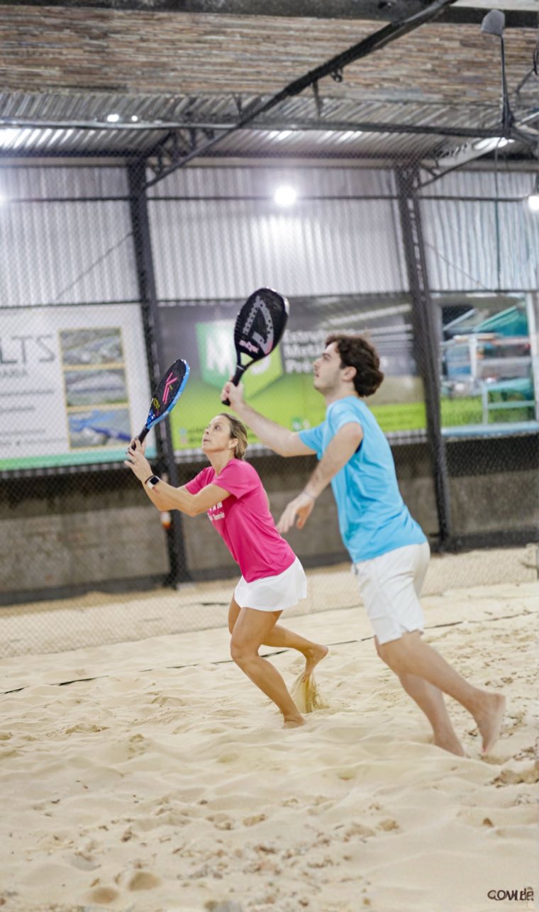 Dupla de beach tennis se preparando para devolver a bola em quadra de areia coberta.